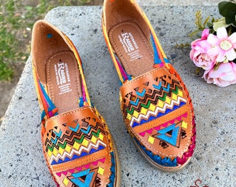 May include: A pair of brown leather sandals with colorful embroidery. The sandals have a zig-zag pattern and a blue, green, yellow, and pink trim. The sandals are made in Mexico by the brand "Raquer" and are labeled "Artesanias Hecho en Mexico".