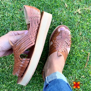 May include: Brown leather sandals with a woven design and a platform sole. The sandals are on a green grassy surface.