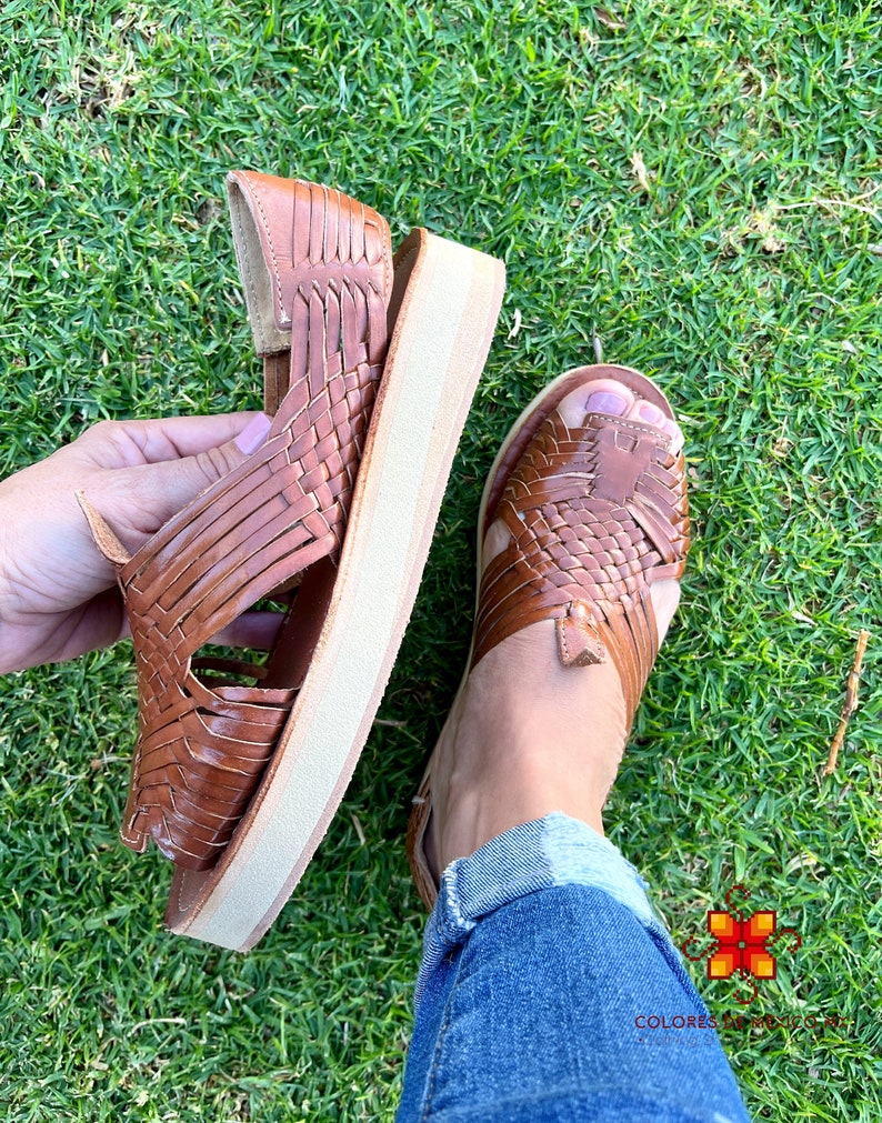 May include: Brown leather sandals with a woven design and a platform sole. The sandals are on a green grassy surface.