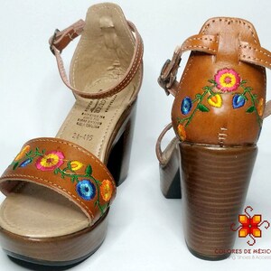 May include: Brown leather sandals with a platform heel and embroidered flowers in shades of pink, yellow, blue, and green. The sandals have an ankle strap with a buckle closure. The text "24-195" is visible on the sole.