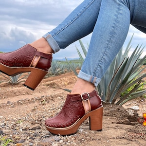 May include: A pair of brown leather ankle boots with a floral pattern and a chunky heel. The boots are worn by a person wearing blue jeans and sitting on a rock.