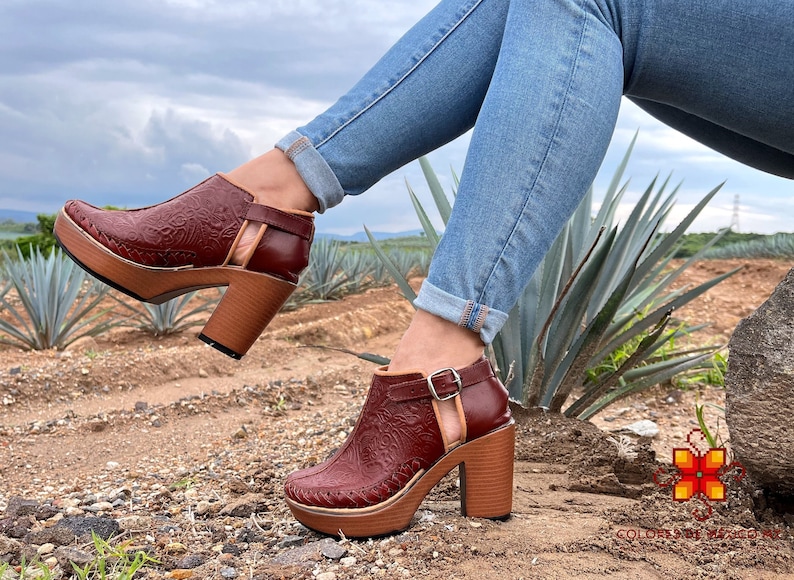May include: A pair of brown leather ankle boots with a floral pattern and a chunky heel. The boots are worn by a person wearing blue jeans and sitting on a rock.