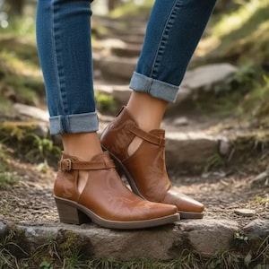 Może przedstawiać: Brązowe skórzane botki z motywem westernowym. Botki mają wycięcie z boku, zapięcie na klamrę i niski obcas. Botki są noszone z niebieskimi dżinsami.