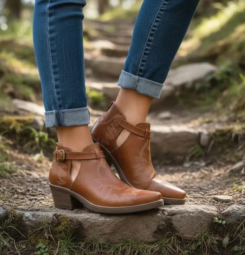 Pu&ograve; includere: Stivaletti in pelle marrone con design di ispirazione western. Gli stivaletti presentano un dettaglio ritagliato sul lato, una chiusura con fibbia e un tacco basso. Gli stivaletti sono indossati con jeans blu.
