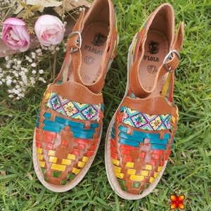 May include: A pair of brown leather huaraches with colorful woven detailing in teal, red, yellow, and green. The sandals have a buckle closure and the brand name "D'Lolos" is visible on the insole. The shoes are set on a white sole.