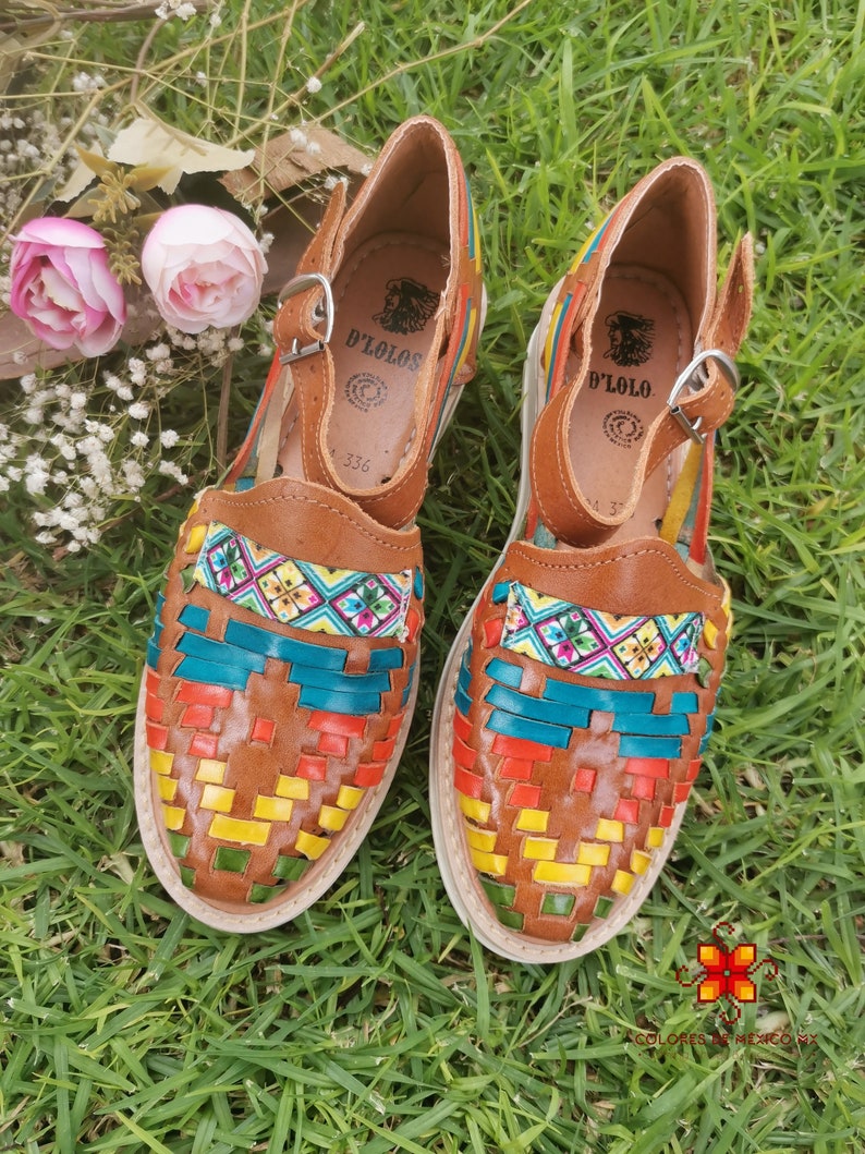 May include: A pair of brown leather huaraches with colorful woven detailing in teal, red, yellow, and green. The sandals have a buckle closure and the brand name "D'Lolos" is visible on the insole. The shoes are set on a white sole.