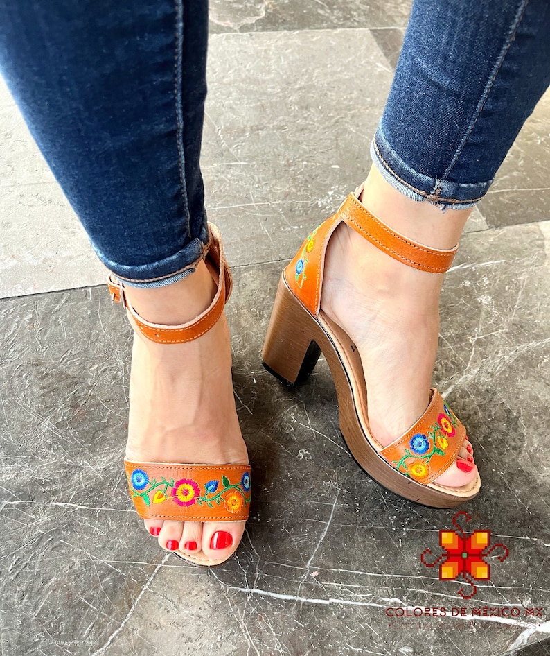 May include: A pair of brown leather heeled sandals with colorful floral embroidery. The sandals have a thick wooden platform and ankle straps.