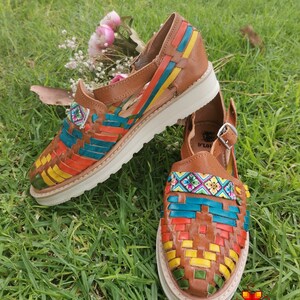 May include: A pair of brown leather huaraches with colorful woven straps in orange, blue, yellow, and green. The shoes have white soles and are displayed on a bed of green grass. A small floral arrangement is in the background.