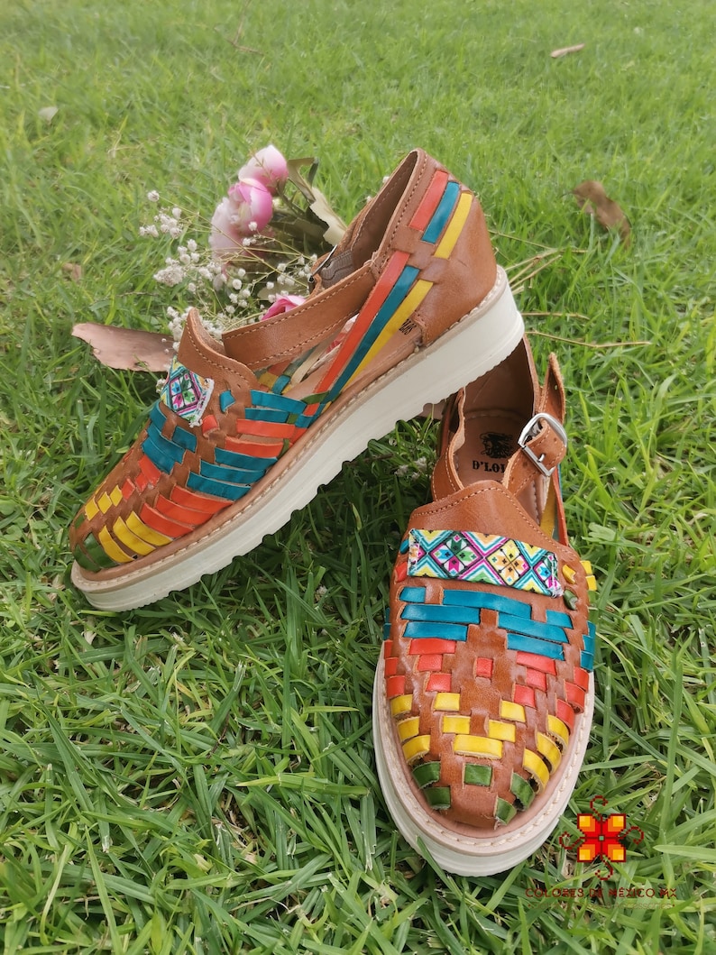 May include: A pair of brown leather huaraches with colorful woven straps in orange, blue, yellow, and green. The shoes have white soles and are displayed on a bed of green grass. A small floral arrangement is in the background.