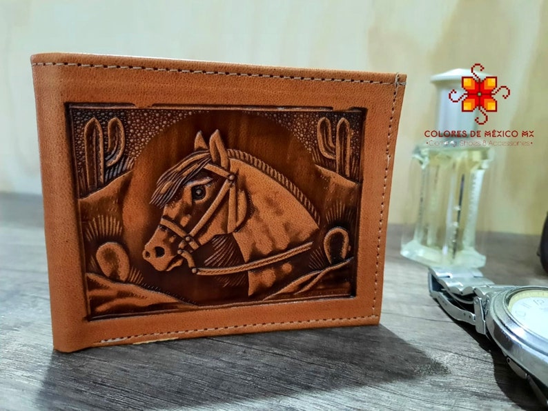 Puede incluir: Una cartera de cuero marr&oacute;n con un dise&ntilde;o estampado de una cabeza de caballo y cactus. La cartera tiene un borde cosido y est&aacute; cerrada.