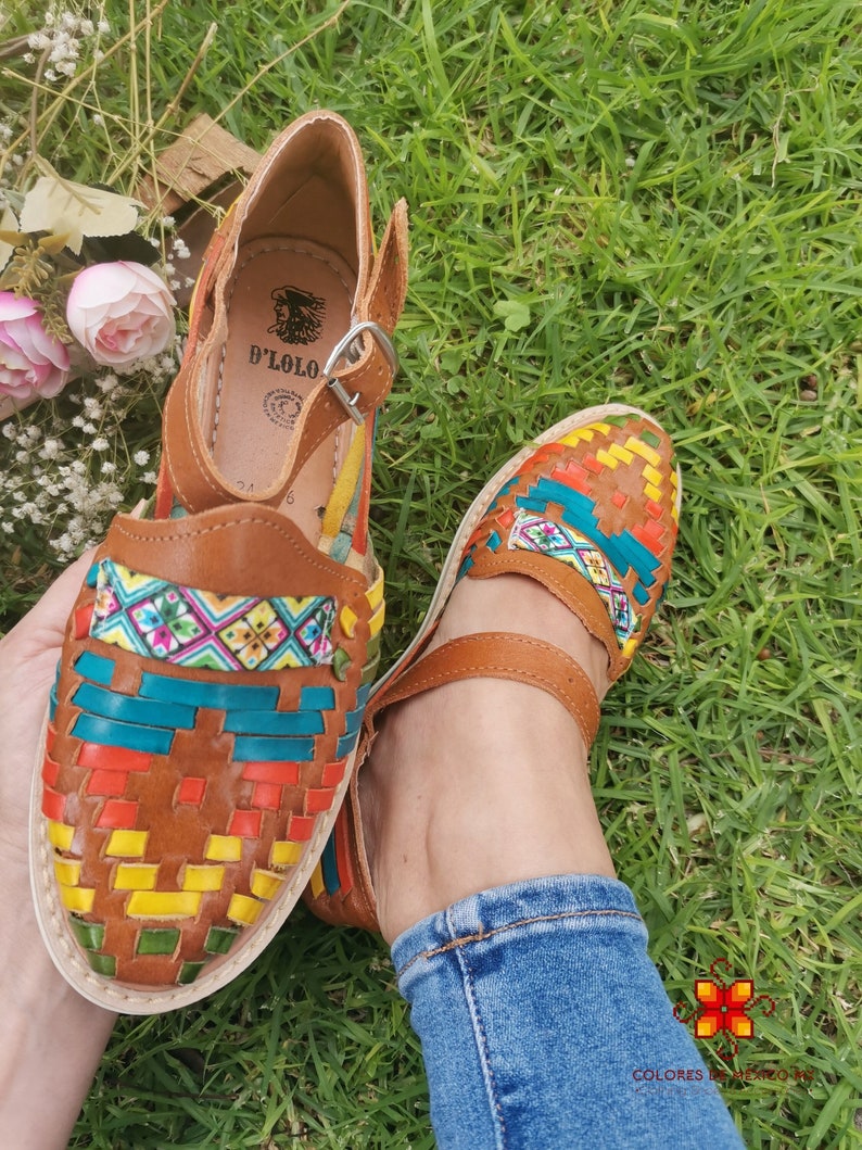 May include: Brown leather sandals with woven colorful accents. The sandals feature a strap and buckle closure, with a multi-colored woven design on the toe and a leather strap. The sandals are on a grassy surface.