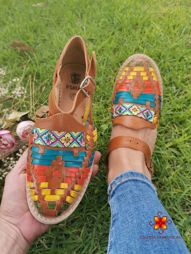 May include: Brown leather sandals with colorful woven detailing. The sandals feature a strap and buckle closure. The woven design incorporates red, yellow, blue, and green hues. The sandals are displayed on a grassy surface.