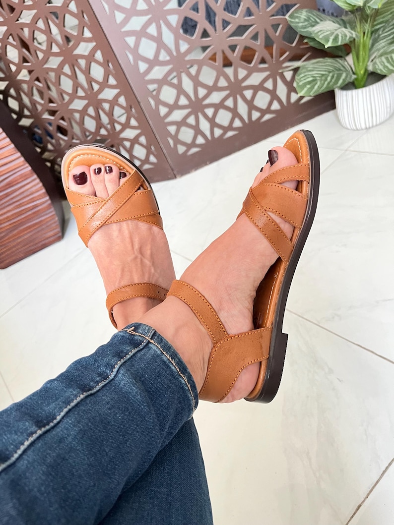 Pu&ograve; includere: Sandali in pelle color cuoio con cinturini incrociati e cinturino alla caviglia. I sandali sono indossati da una persona che indossa jeans blu.