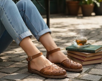 Puede incluir: Un par de sandalias huarache de cuero marrón con un diseño superior trenzado, punta cerrada y una correa de tobillo ajustable con hebilla de metal. Las sandalias se usan con jeans de mezclilla azul claro con el dobladillo vuelto.