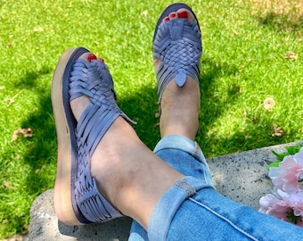 May include: A pair of gray woven leather sandals with a platform sole. The sandals are worn by a person sitting on a concrete surface with green grass in the background.