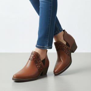 Może przedstawiać: Brązowe skórzane botki z dekoracyjnym kwiatowym wzorem z boku. Botki mają spiczasty nosek i obcas w kształcie klocka. Osoba ma na sobie niebieskie dżinsy. Botki są w ciepłym brązowym kolorze. Obcas ma około 5 cm wysokości.