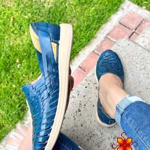 May include: A pair of blue leather sandals with woven straps. The sandals have a white sole and are being worn by a person sitting on a concrete step.