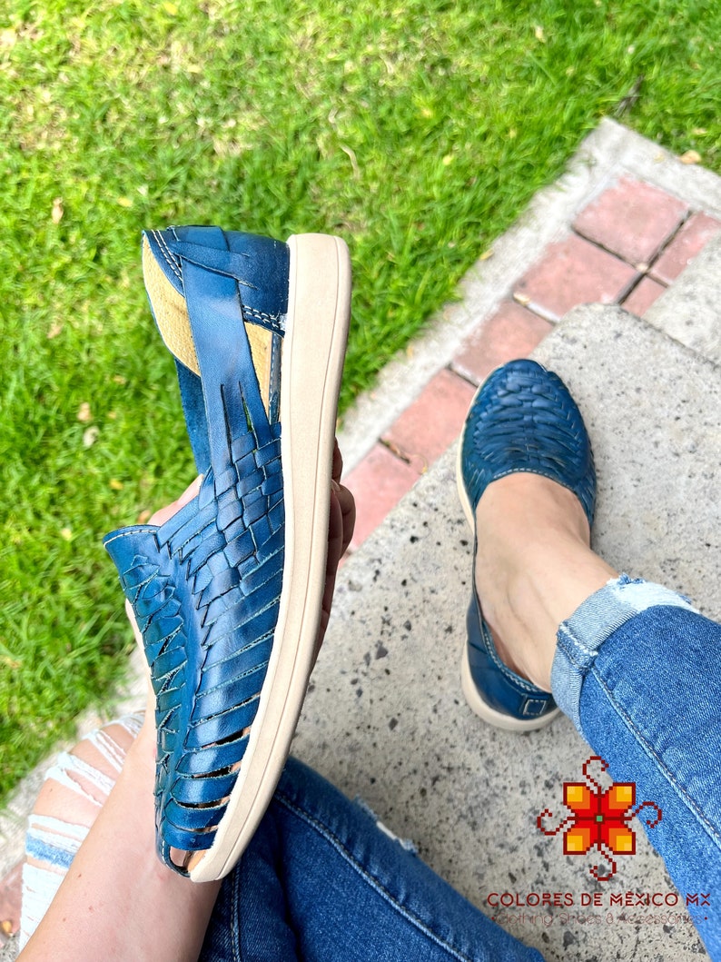 May include: A pair of blue leather sandals with woven straps. The sandals have a white sole and are being worn by a person sitting on a concrete step.