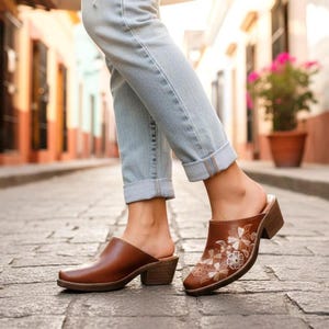 Puede incluir: Zuecos de cuero marrón con un diseño floral en la parte superior. Los zuecos tienen un tacón de bloque bajo y un diseño sin cordones. La persona de la imagen lleva vaqueros azul claro. Los zuecos son de estilo clásico.