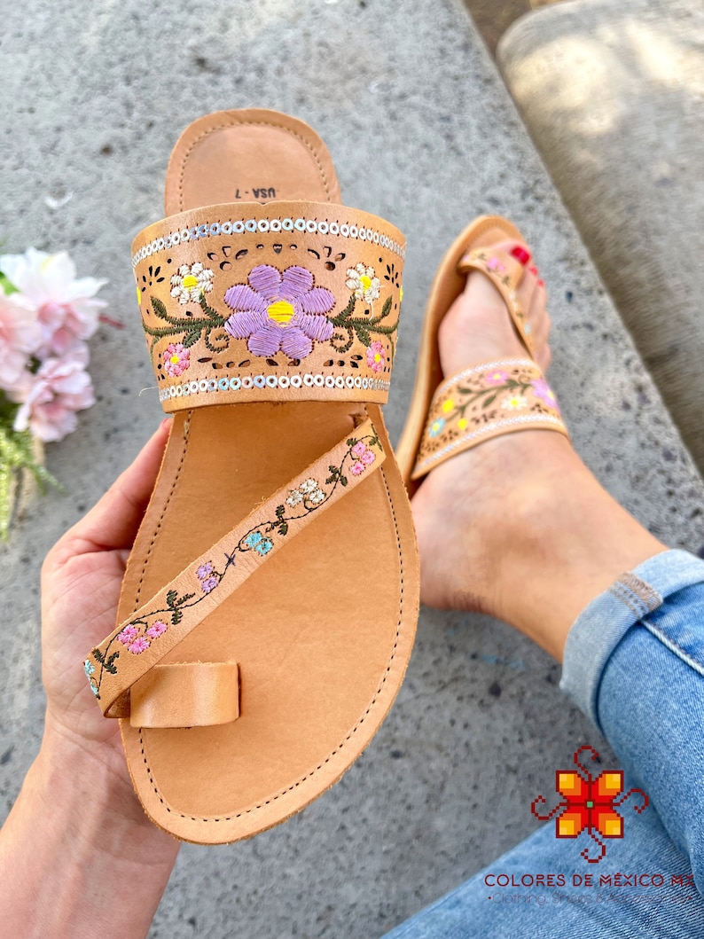 K&ouml;nnte beinhalten: Tanfarbene Ledersandalen mit Blumenstickerei. Die Sandalen haben eine Zehenschlaufe und einen Kn&ouml;chelriemen. Die Stickerei ist in den Farben Rosa, Lila, Gelb und Gr&uuml;n gehalten.
