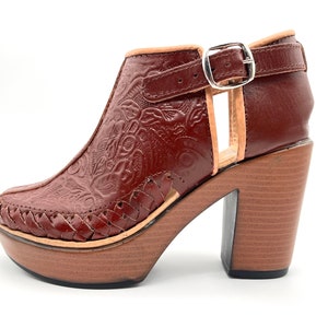 May include: Brown leather heeled ankle boots with a floral embossed design and a woven leather detail around the ankle. The boots have a chunky wooden heel and a buckle closure.
