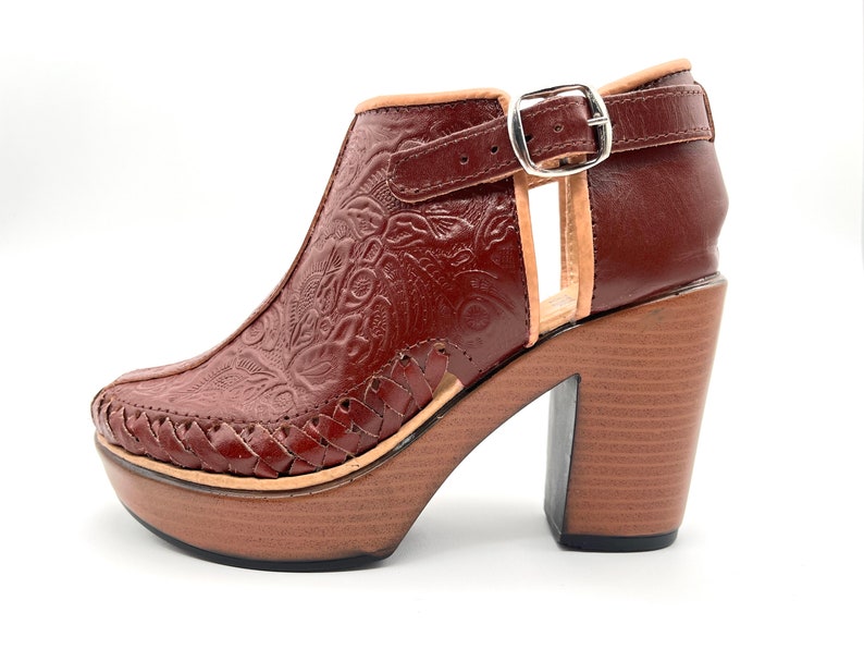 May include: Brown leather heeled ankle boots with a floral embossed design and a woven leather detail around the ankle. The boots have a chunky wooden heel and a buckle closure.