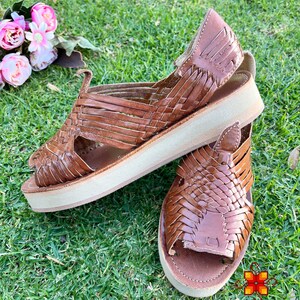 May include: A pair of brown leather sandals with woven straps and a platform sole. The sandals are on a green grassy surface.