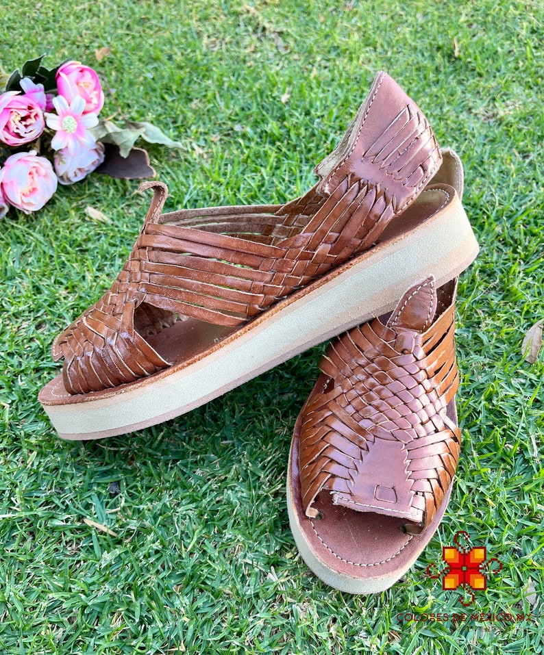 May include: A pair of brown leather sandals with woven straps and a platform sole. The sandals are on a green grassy surface.