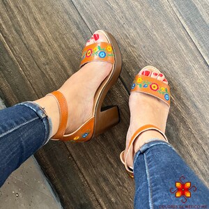 May include: Brown leather sandals with a chunky wooden heel and colorful floral embroidery. The sandals have an ankle strap and are worn with blue jeans.