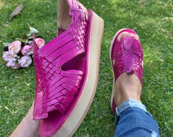 May include: A pair of bright pink woven leather sandals with a platform sole. The sandals are worn by a person sitting on the grass.