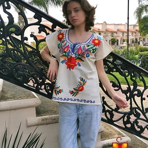 May include: A white embroidered blouse with colorful floral designs. The blouse has a scoop neck and short sleeves. The model is wearing blue jeans and standing on a set of stairs.