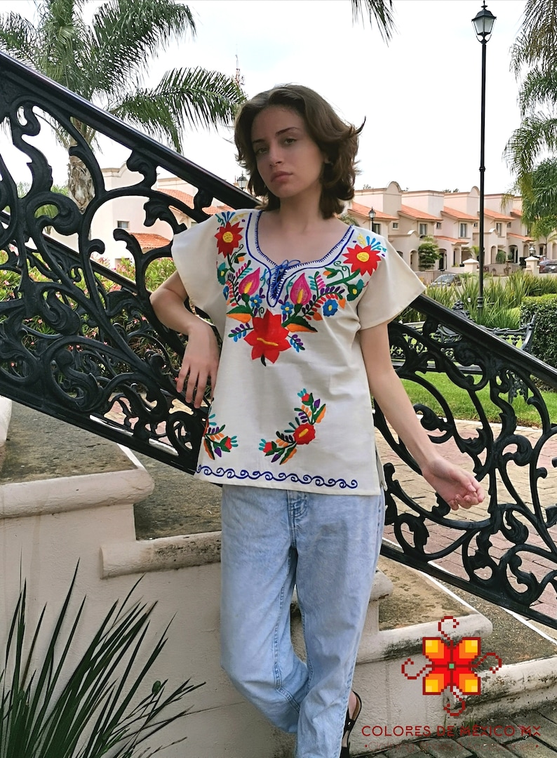 May include: A white embroidered blouse with colorful floral designs. The blouse has a scoop neck and short sleeves. The model is wearing blue jeans and standing on a set of stairs.