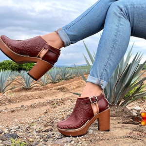 May include: A pair of brown leather heeled shoes with intricate floral designs. The shoes have a wooden heel and a cut-out design on the side. The shoes are worn by a person wearing blue jeans and are photographed in a field of agave plants.
