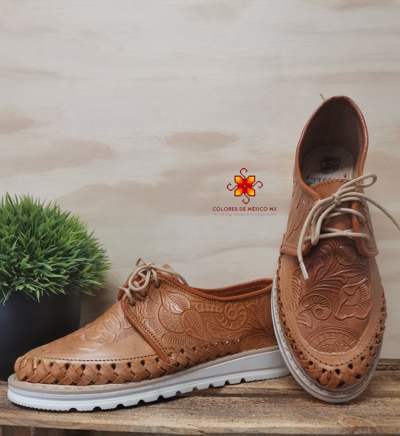 K&ouml;nnte beinhalten: Ein Paar braune Lederschuhe mit aufwendigen Blumenstickereien. Die Schuhe haben eine wei&szlig;e Sohle und sind mit wei&szlig;en Schn&uuml;rsenkeln gebunden. Die Schuhe befinden sich auf einer Holzoberfl&auml;che.