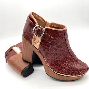May include: A pair of burgundy leather heeled shoes with a floral embossed design. The shoes have a buckle strap and a wooden platform sole. The brand name "SOLOS" is visible on the side of the shoe.