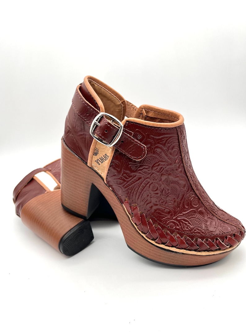 May include: A pair of burgundy leather heeled shoes with a floral embossed design. The shoes have a buckle strap and a wooden platform sole. The brand name "SOLOS" is visible on the side of the shoe.