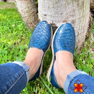 May include: A pair of blue leather sandals with a woven design. The sandals are being worn by a person sitting on the grass.