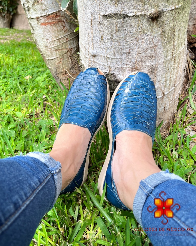 May include: A pair of blue leather sandals with a woven design. The sandals are being worn by a person sitting on the grass.