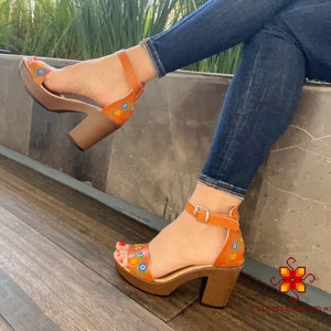 May include: A pair of brown leather sandals with a chunky heel and floral embroidery. The sandals have a strap across the ankle and a buckle closure.