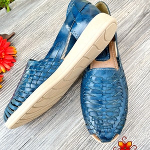 May include: A pair of blue leather sandals with woven straps and a white sole. The sandals are on a wooden surface.