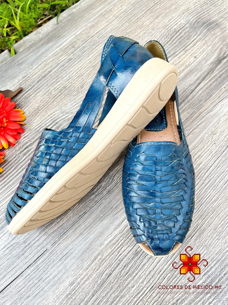 May include: A pair of blue leather sandals with woven straps and a white sole. The sandals are on a wooden surface.