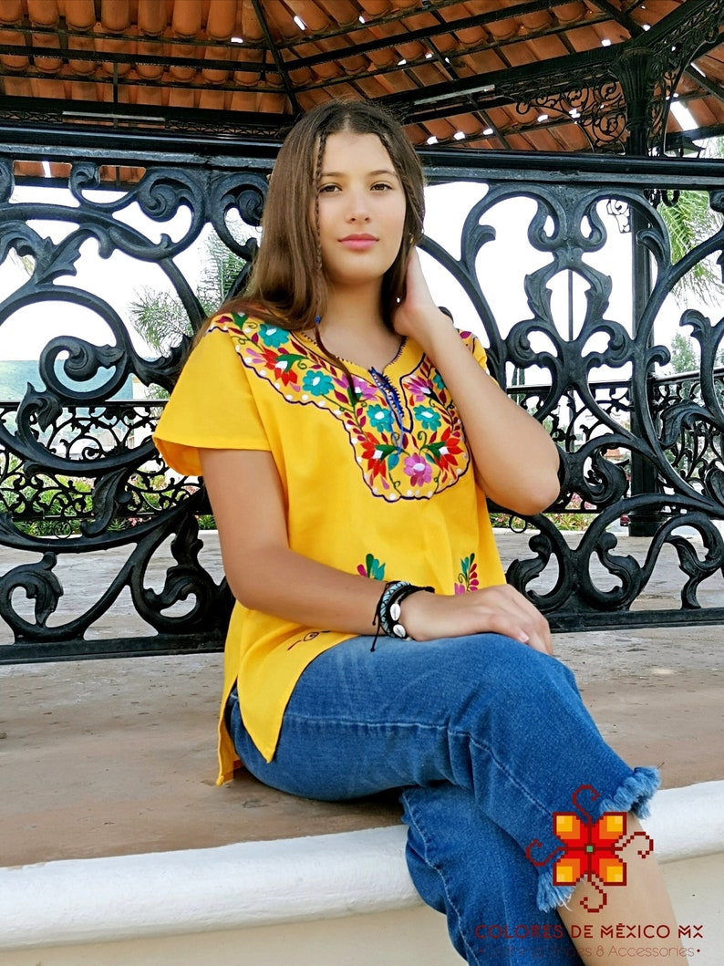May include: A yellow embroidered blouse with colorful floral and geometric patterns. The blouse has short sleeves and a V-neckline. The model is wearing blue jeans and sitting on a bench.