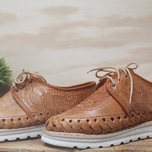 K&ouml;nnte beinhalten: Ein Paar braune Lederschuhe mit aufwendigen Blumenmustern und gewebten Details an den Sohlen. Die Schuhe haben wei&szlig;e Schn&uuml;rsenkel und wei&szlig;e Sohlen. Die Schuhe werden von "Colores de Mexico MX" hergestellt.