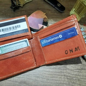 Puede incluir: Una cartera de cuero marr&oacute;n con una ranura para tarjeta de cr&eacute;dito y un portafolios. La cartera tiene una inscripci&oacute;n personalizada que dice "OHM".