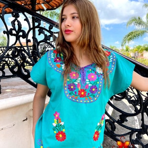 May include: A turquoise blouse with colorful floral embroidery. The embroidery features flowers in shades of pink, red, yellow, and white. The blouse has short sleeves and a round neckline.
