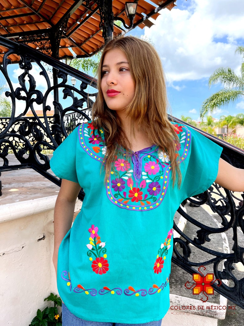 May include: A turquoise blouse with colorful floral embroidery. The embroidery features flowers in shades of pink, red, yellow, and white. The blouse has short sleeves and a round neckline.