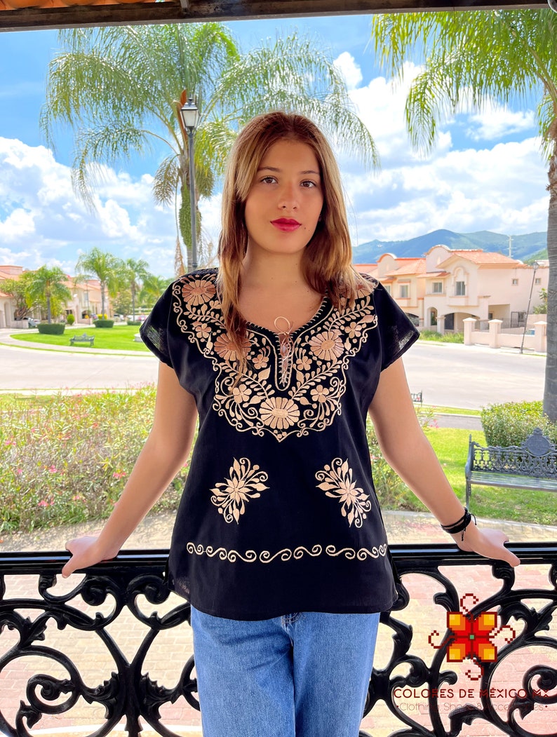 May include: A black embroidered blouse with gold floral designs. The blouse has short sleeves and a round neckline. The model is wearing blue jeans.