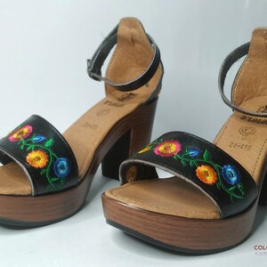 May include: Black leather sandals with a floral embroidery design. The sandals have a wooden platform and a high heel. The brand name "D'ZOLOS" is visible on the sandals.