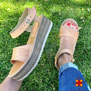Peut inclure: Une paire de sandales en cuir marron clair avec un motif floral. Les sandales ont une &eacute;paisse semelle plateforme et une sangle de cheville r&eacute;glable avec une boucle. Les sandales sont port&eacute;es par une personne avec du vernis &agrave; ongles rouge sur les orteils.