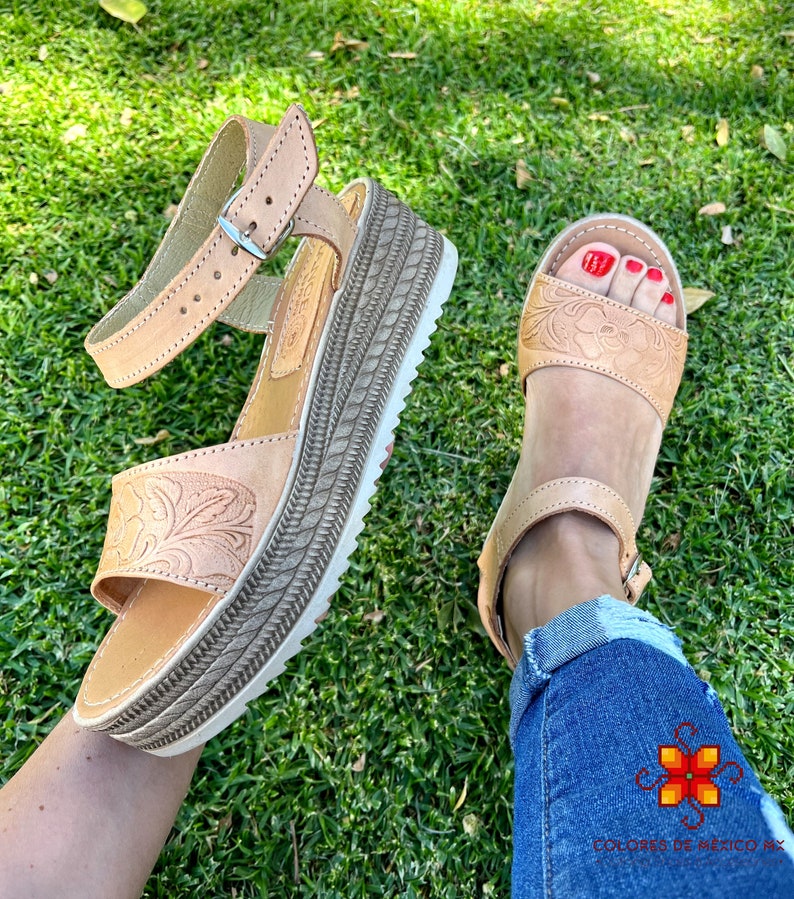 Peut inclure: Une paire de sandales en cuir marron clair avec un motif floral. Les sandales ont une &eacute;paisse semelle plateforme et une sangle de cheville r&eacute;glable avec une boucle. Les sandales sont port&eacute;es par une personne avec du vernis &agrave; ongles rouge sur les orteils.